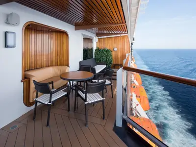 Sonniger Balkon einer Suite an Bord der Westerdam mit Sitzgruppe, gepolstertem Sofa, Holzverkleidung und Blick auf das Meer.