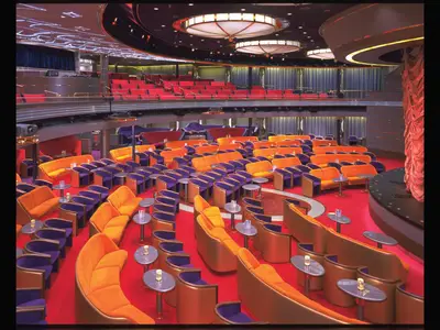 Die mehrstöckige Show-Lounge an Bord der Zaandam mit roten Teppichen, orangefarbenen Polstersesseln und einer Bühne am rechten Rand.