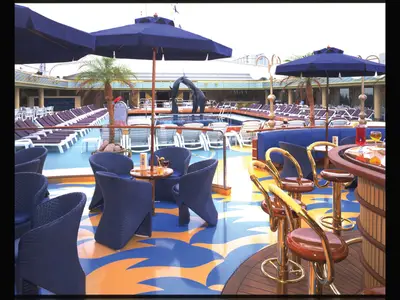Außenbereich an Bord der Zaandam mit Blick auf den Lido-Pool, eine Delfinskulptur, blaue Sonnenschirme, Liegestühle und gemütliche Bar-Sitzgruppen.