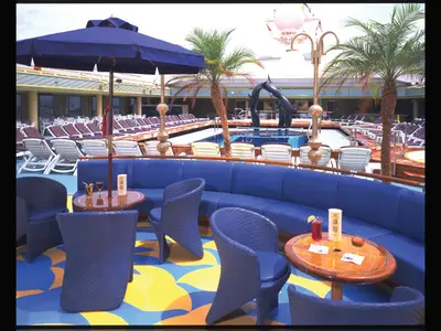 Blaue Sessel und Holzbeistelltische mit Cocktails im Loungebereich an Bord der Zaandam, im Hintergrund der Lido Pool.