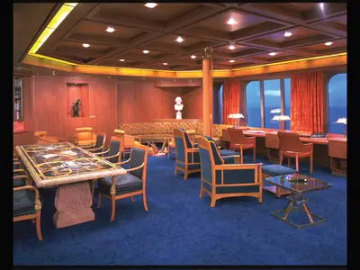 Ein Blick in die gemütliche Bibliothek an Bord der Zaandam mit blauer Auslegeware, Holzmöbeln und Fenstern zum Meer.