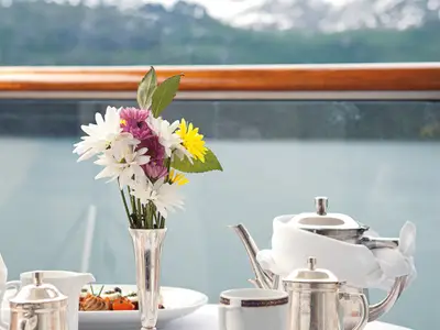 Ein gedeckter Tisch mit bunten Blumen in einer Vase und einer Teekanne auf der Veranda an Bord der Zuiderdam vor einer Berglandschaft.