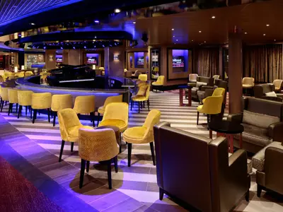 Eine elegante Lounge an Bord der Zuiderdam mit gelben Sesseln, bequemen Sesseln, einer Bar und einem Klavier in der Mitte.