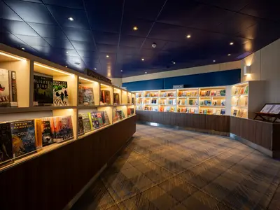 Blick in die moderne und gemütlich beleuchtete Bibliothek an Bord der Zuiderdam mit zahlreichen Buchregalen und Teppichboden.