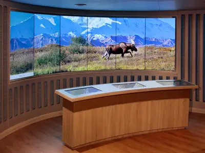 An Bord der Zuiderdam: Ein Informationsbereich mit interaktiven Tisch-Bildschirmen und einer großen Videowand, die eine Landschaft in Alaska zeigt.