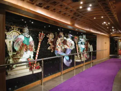 Ein elegant beleuchteter Gang an Bord der Zuiderdam mit einem lila Teppich und kunstvollen venezianischen Figuren hinter Glas.
