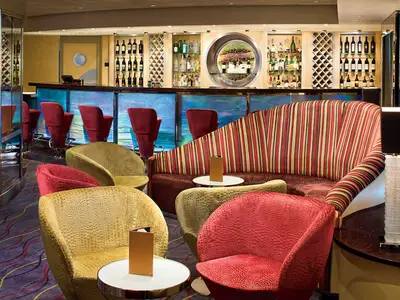 Stilvolle Bar an Bord der Zuiderdam mit gemusterten Sesseln in Rot und Gold, einem gestreiften Sofa und einer beleuchteten Barzeile.