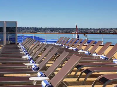 Reihen von braunen Liegestühlen mit blau-weiß gestreiften Handtüchern auf dem Sonnendeck an Bord der Zuiderdam vor einer Küstenstadt.