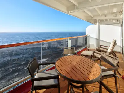 Balkon einer Neptune Suite an Bord der Zuiderdam mit Holztisch, Stühlen, Liegen und weitem Blick aufs blaue Meer.