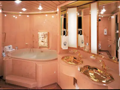 Luxuriöses Badezimmer der Pinnacle Suite an Bord der Zuiderdam mit runder Badewanne, goldenen Waschbecken und Armaturen sowie rosa Marmor.