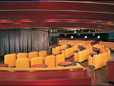 Blick in die Queen's Lounge an Bord der Zuiderdam mit eleganten orangefarbenen Sesseln vor einer Bühne mit glitzerndem Vorhang.