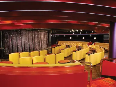 Blick in die stilvolle Queen's Lounge an Bord der Zuiderdam mit gelben Sesseln, kleinen Tischen und einer glitzernden Showbühne.
