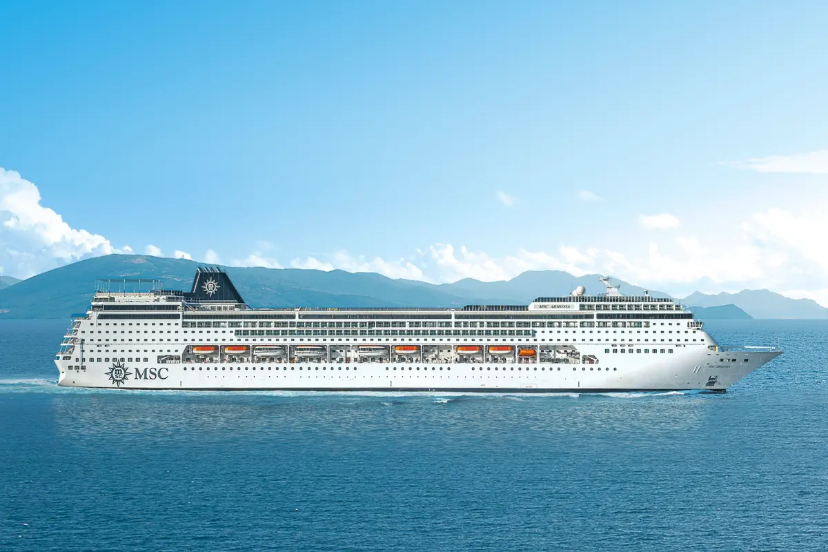 Ein großes weißes Kreuzfahrtschiff, die MSC Armonia, fährt auf blauem Meer. Im Hintergrund sind grüne Berge und ein klarer Himmel mit Wolken zu sehen. Das Schiff hat ein schwarzes Schornsteinlogo und das MSC-Logo am Heck.
