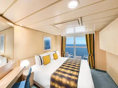Innenansicht einer hellen Balkonkabine der MSC Armonia mit einem Doppelbett, gelben Kissen und einer karierten Decke. Ein großer Spiegel und ein Schreibtisch sind links zu sehen. Durch die gläserne Balkontür ist das blaue Meer mit zwei Stühlen auf dem Balkon sichtbar.
