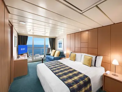 Innenansicht einer geräumigen Kabine der MSC Armonia. Ein großes Bett mit karierter Tagesdecke und gelben Kissen dominiert den Vordergrund, daneben ein Sofa und ein TV mit MSC Logo. Eine Glastür führt zu einem Balkon mit Blick auf das blaue Meer.