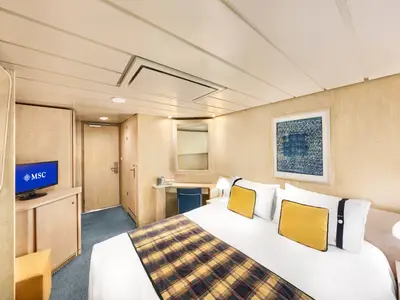 Innenansicht einer hellen Kreuzfahrtkabine mit Doppelbett, plaidfarbener Tagesdecke, gelben Kissen, einem TV mit MSC Logo und einem Schminktisch mit Spiegel und blauem Stuhl.