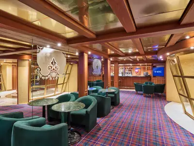 Innenansicht einer Bar-Lounge an Bord der MSC Armonia mit grün-blau kariertem Teppich, dunklen Holzdeckenbalken und hellgoldenen Deckenelementen. Mehrere dunkelgrüne Sessel und kleine Marmortische stehen auf dem Teppich. Im Hintergrund befindet sich eine Bar mit Holztheke und ein Bildschirm mit dem MSC Logo.