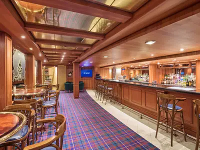 Innenansicht der Bar-Lounge der MSC Armonia mit einer langen, gut bestückten Bar auf der rechten Seite und mehreren Bartischen und Stühlen auf der linken Seite. Der Boden ist mit einem karierten Teppich ausgelegt, der in Blau-, Rot- und Grüntönen gehalten ist. Die Wände und die Decke sind mit hellem Holz verkleidet. Im Hintergrund sind zwei dunkle Sessel und ein Fernseher mit dem MSC-Logo zu sehen.