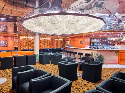 Blick in die schicke Bar-Lounge der MSC Armonia mit einer langen, halbrunden Bar auf der rechten Seite. Im Vordergrund sind viele schwarze Sessel um kleine Tische gruppiert. Der Boden ist mit einem orange-schwarz gemusterten Teppich ausgelegt. Eine orangefarbene Sitzbank schlängelt sich im Hintergrund, umrahmt von gemusterten Vorhängen und Wänden. Die Decke ist spiegelnd. Ein gigantischer Kronleuchter überragt den Raum.