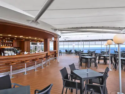 Eine große, geschwungene Holzbar mit Barhockern und einer Espresso-Maschine an Bord der MSC Armonia. Davor erstreckt sich ein weitläufiger Außenbereich mit zahlreichen dunklen Tischen und Stühlen, beleuchtet von mehreren runden Stehlampen. Im Hintergrund ist das blaue Meer zu sehen, eingerahmt von einer Reling.