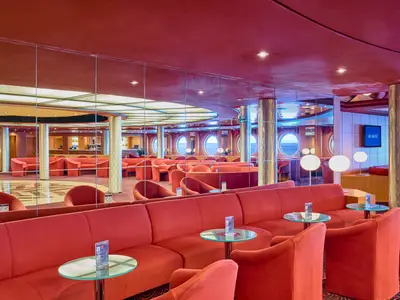 Ein weiter Blick auf die Lounge an Bord der MSC Armonia, die mit vielen roten Polstermöbeln, runden Glastischen und goldenen Säulen ausgestattet ist. Große Bullaugen ermöglichen einen Blick auf das Meer. Über Spiegel an der Decke und an den Wänden wird der Raum zusätzlich erweitert.