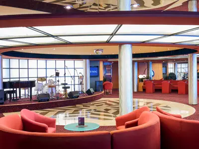 Innenansicht der Bar-Lounge an Bord der MSC Armonia, mit einer Bühne, auf der ein Flügel und ein Schlagzeug stehen. Zahlreiche rote Sessel und Sofas sind auf einem roten Teppichboden arrangiert, während im Zentrum des Raumes ein gemusterter Fliesenboden zu sehen ist.