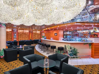 Blick in die schicke Lounge und Bar an Bord der MSC Armonia. Eine helle Bar mit tropischen Motiven zieht sich entlang der rechten Seite. Davor stehen Barhocker und bequeme schwarze Sessel mit Glastischen. Die Decke ist mit einer großen, funkelnden Kristallwolke bedeckt.