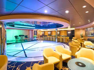 Blick in die Armonia Lounge eines Kreuzfahrtschiffs mit heller Tanzfläche in der Mitte, umgeben von gelben Sesseln. Im Hintergrund eine kleine Bühne mit Stühlen und eine Bar.