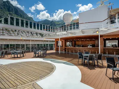 Blick von oben auf das Deck der MSC Armonia mit einer gut ausgestatteten Holzbar, Tischen und Stühlen. Im Hintergrund sind grüne Berge und ein blauer Himmel zu sehen.