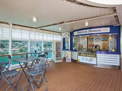 Innenansicht der Gelateria Italiana an Bord der MSC Armonia mit einer Eisdiele und mehreren Tischen und Stühlen. Durch große Fenster ist eine grüne Küste mit vielen Häusern und das türkisfarbene Meer zu sehen.