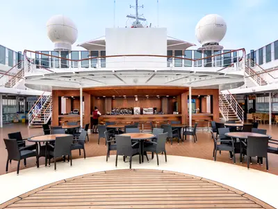 Blick auf die Poolbar und einen Teil des Außendecks der MSC Armonia. Eine Barkeeperin steht hinter der Bar, umgeben von dunklen Tischen und Stühlen. Im Hintergrund sind Treppen und ein Gelato-Stand zu sehen.