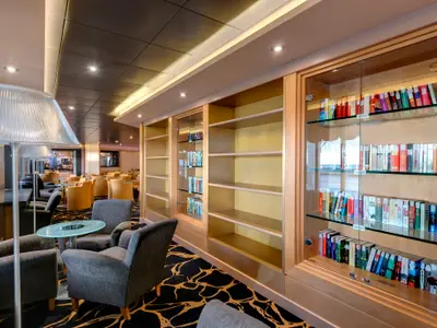 Blick in die elegante Lese-Lounge der MSC Armonia mit bequemen Polstersesseln, runden Tischen und deckenhohen, hellholzfarbenen Bücherregalen. Einige Regale sind mit Glasfronten und Büchern gefüllt, andere sind leer. Ein gläserner Stehlampenschirm im Vordergrund.
