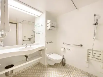 Ein helles, weißes, barrierefreies Badezimmer an Bord der MSC Armonia. Es verfügt über eine große Spüle, einen Spiegel, eine Toilette mit Haltegriffen, einen Duschbereich mit Duschkopf und einen klappbaren Duschsitz.