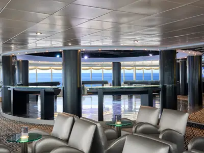 Blick in eine elegante Bar an Bord der MSC Armonia mit großen Fenstern zum Meer. Zahlreiche Sessel sind um runde Tische gruppiert. Im Hintergrund sind dunkle Säulen und eine Bar zu sehen.
