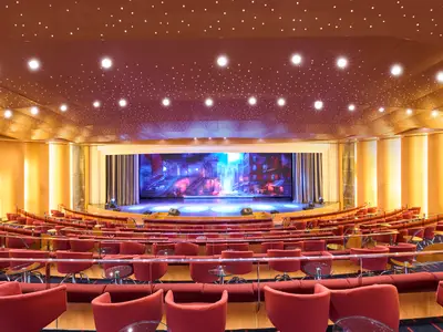 Blick in das geräumige Theater der MSC Armonia mit Reihen von roten Sesseln und kleinen Tischen. Die Decke ist dunkel mit vielen kleinen Lichtern, die einen Sternenhimmel simulieren. Die Bühne zeigt eine farbenfrohe Stadtlandschaft auf einem großen Bildschirm.