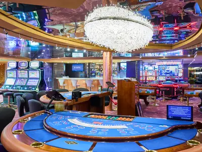 Ein luxuriöses Casino an Bord der MSC Armonia, das mit glänzenden Spielautomaten, blauen Kartentischen und einer gut bestückten Bar ausgestattet ist. An der Decke hängt ein großer, auffälliger Kronleuchter.