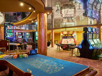 Blick in das beleuchtete Casino an Bord der MSC Armonia. Im Vordergrund ein blauer Roulettetisch mit bunten Chips. Im Hintergrund sind zahlreiche leuchtende Spielautomaten und ein "Las Vegas"-Automat zu sehen. Das Interieur ist geprägt von warmen Holztönen, goldenen Säulen und reflektierenden Decken.