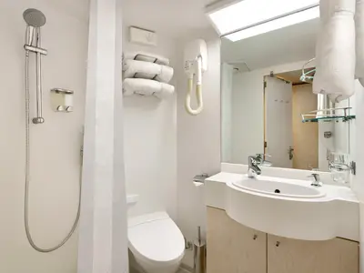 Dieses Foto zeigt ein kleines, hell eingerichtetes Badezimmer einer Innenkabine an Bord der MSC Armonia. Links ist eine Duschkabine mit verstellbarer Duschstange und Spender. In der Mitte befindet sich eine Toilette und rechts ein Waschbecken mit Armatur, Seifenspender und einem Spiegel darüber. Am Spiegel hängen zwei weiße Handtücher und darunter sind Gläser in einer Halterung zu sehen. An der Wand hängen außerdem aufgerollte Handtücher und ein Föhn.