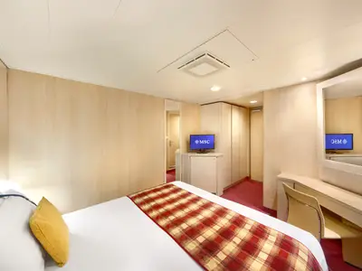 Ein helles Schlafzimmer in einer Innenkabine der MSC Armonia, ausgestattet mit einem großen Bett, einem Fernseher auf einem Holzunterschrank und einem Schminktisch mit Stuhl. Eine Deckenleuchte und ein Lüftungsgitter sind an der Decke sichtbar. Der Boden ist mit einem roten Teppich ausgelegt.