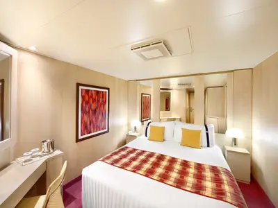 Blick in eine freundliche Innenkabine der MSC Armonia mit einem großen Doppelbett, zwei Nachttischen mit Lampen und einem Schreibtisch mit Spiegel auf der linken Seite. Ein farbenfrohes Kunstwerk hängt über dem Bett. Helle Holzwände und ein karierter Bettläufer in Rot- und Gelbtönen prägen den Raum.