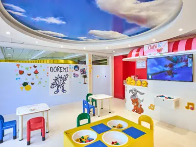 Ein hell erleuchteter Kinderbereich an Bord der MSC Armonia mit einem Wolkenhimmel-Deckengemälde. Links sind kleine Tische und Stühle zu sehen, rechts ein "Café"-Bereich mit Fernseher und Tierszenen an der Wand.