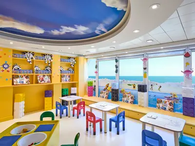 Blick in den fröhlichen Kinderbereich auf der MSC Armonia. Mehrere Tische mit Stühlen stehen vor großen Fenstern, die den Blick auf das blaue Meer freigeben. Die Wände sind gelb und mit Plüschtieren sowie Nintendo Labo-Spielen dekoriert. Bunte Bausteine und eine Himmelsdecke runden das kinderfreundliche Ambiente ab.