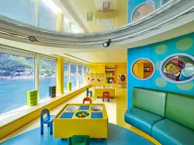 Blick in den farbenfrohen Kinderbereich der MSC Armonia mit großen Fenstern zum Meer, Spieltischen mit Legosteinen und einer grünen Sitzbank. An den Wänden sind LEGO-Figuren als Dekoration zu sehen.