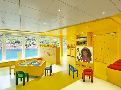 Ein sonniger, gelb gestrichener Innenraum an Bord der MSC Armonia, der als Kinderbereich dient. Große Fenster bieten einen Ausblick auf grüne Hügel und blaues Meer. Im Vordergrund steht ein gelber Spieltisch mit Legobausteinen und kleinen bunten Stühlen. Im Raum sind weitere gelbe Möbel und Dekorationen im Lego-Stil zu sehen, darunter eine Wand mit Fruchtmotiven und ein Mosaikbild.