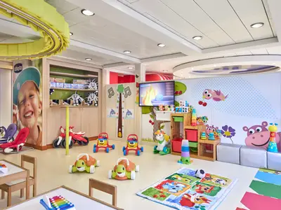 Ein geräumiger, heller Kinderbereich auf der MSC Armonia mit gelbem Boden und weißem Deckenlicht. An den Wänden sind fröhliche Tiere wie Vögel und Nilpferde gemalt. Verschiedene Spielzeuge wie Lauflernhilfen in Schildkrötenform, kleine rote Motorräder und Bauklötze sind im Raum verteilt. Eine Leinwand zeigt Zeichentrickfilme. Im Hintergrund ein Regal mit weiteren Kinderspielzeugen.