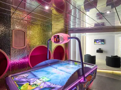 Ein farbenfroher Raum an Bord der MSC Armonia mit einem lila Airhockey-Tisch im Vordergrund. Die Wände sind mit glänzenden, strukturierten Paneelen in Gold und Silber verkleidet und mit pinkfarbenen Lichteinflüssen versehen. Im Hintergrund befindet sich eine weiße Lounge mit zwei schwarzen Sesseln und einem an der Wand montierten Fernseher, auf dem ein Videospiel läuft.