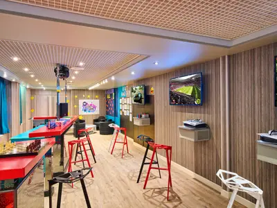 Ein lebendiger Kinderbereich an Bord der MSC Armonia mit einer langen, roten Theke, Schach und Brettspielen. Mehrere Bildschirme zeigen Videospiele. Bunte Hocker und Wanddekorationen mit Emojis und Sternen sind zu sehen.