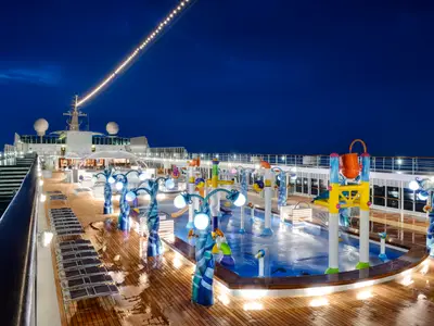 Blick von oben auf den beleuchteten Aquapark der MSC Armonia bei Nacht. Bunte Wasserspiele mit Eimer und Sprinklern, umgeben von einem Holzboden mit Liegestühlen. Im Hintergrund ist das obere Deck des Schiffs zu sehen.