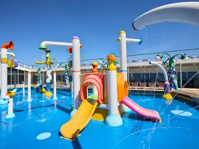 Blick auf den farbenfrohen Wasserpark der MSC Armonia mit blauen Pools, gelben und pinken Rutschen, sowie diversen Wasserspielen unter strahlend blauem Himmel. Im Hintergrund sind Liegestühle und andere Decks des Schiffs zu sehen.