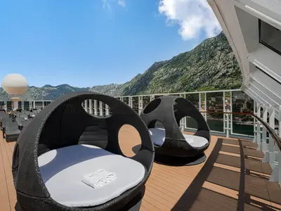 Ein sonniges Deck der MSC Armonia mit zwei stilvollen, schwarzen Rattan-Liegeinseln und vielen Liegestühlen im Hintergrund. Im Hintergrund sind steile, grüne Berge und eine Stadt am Wasser zu sehen, überragt von einem blauen Himmel mit ein paar Wolken.